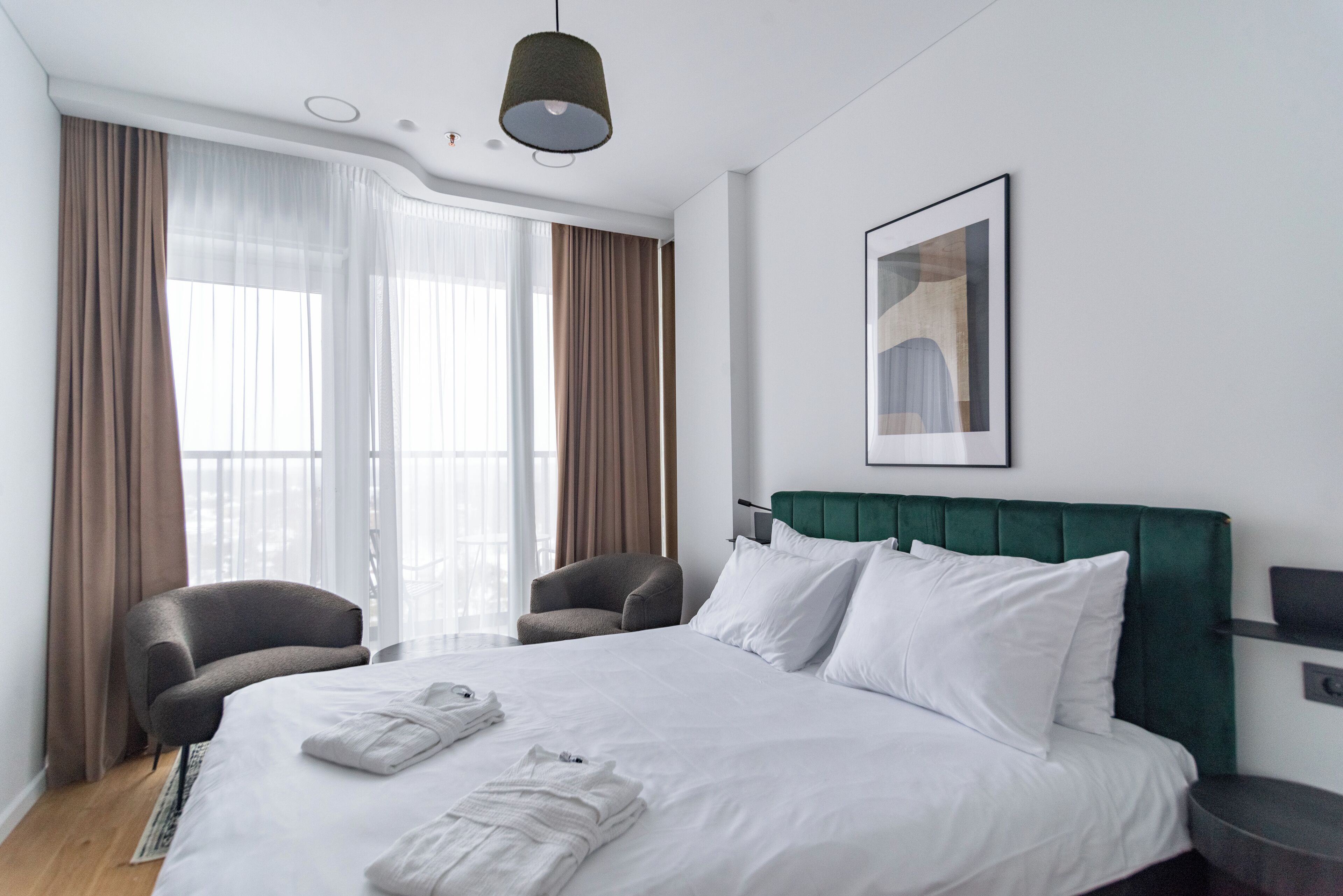 Quarto de hotel com uma cama grande, duas poltronas, cabeceira verde e um quadro na parede.