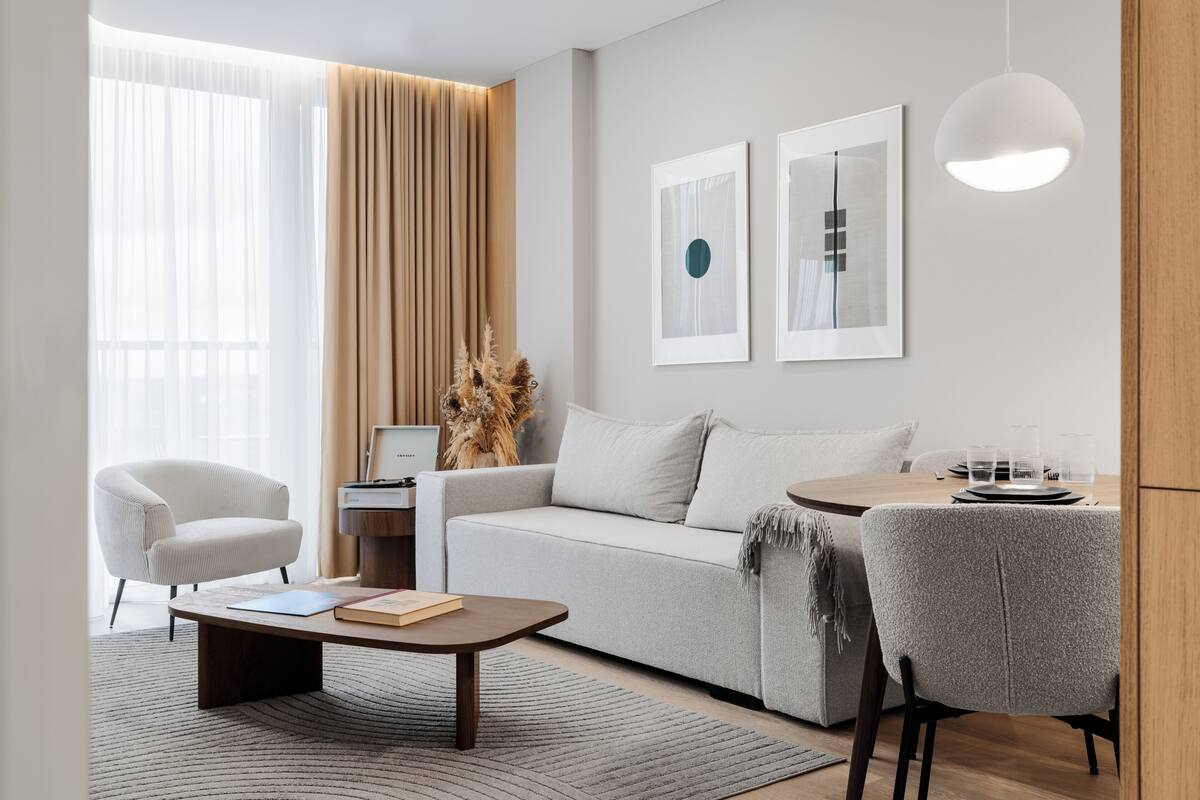 Een moderne woonkamer met een bank, fauteuils, een salontafel en een eettafel met stoelen. Twee ingelijste foto's hangen aan de muur en er staat een lamp op de tafel.