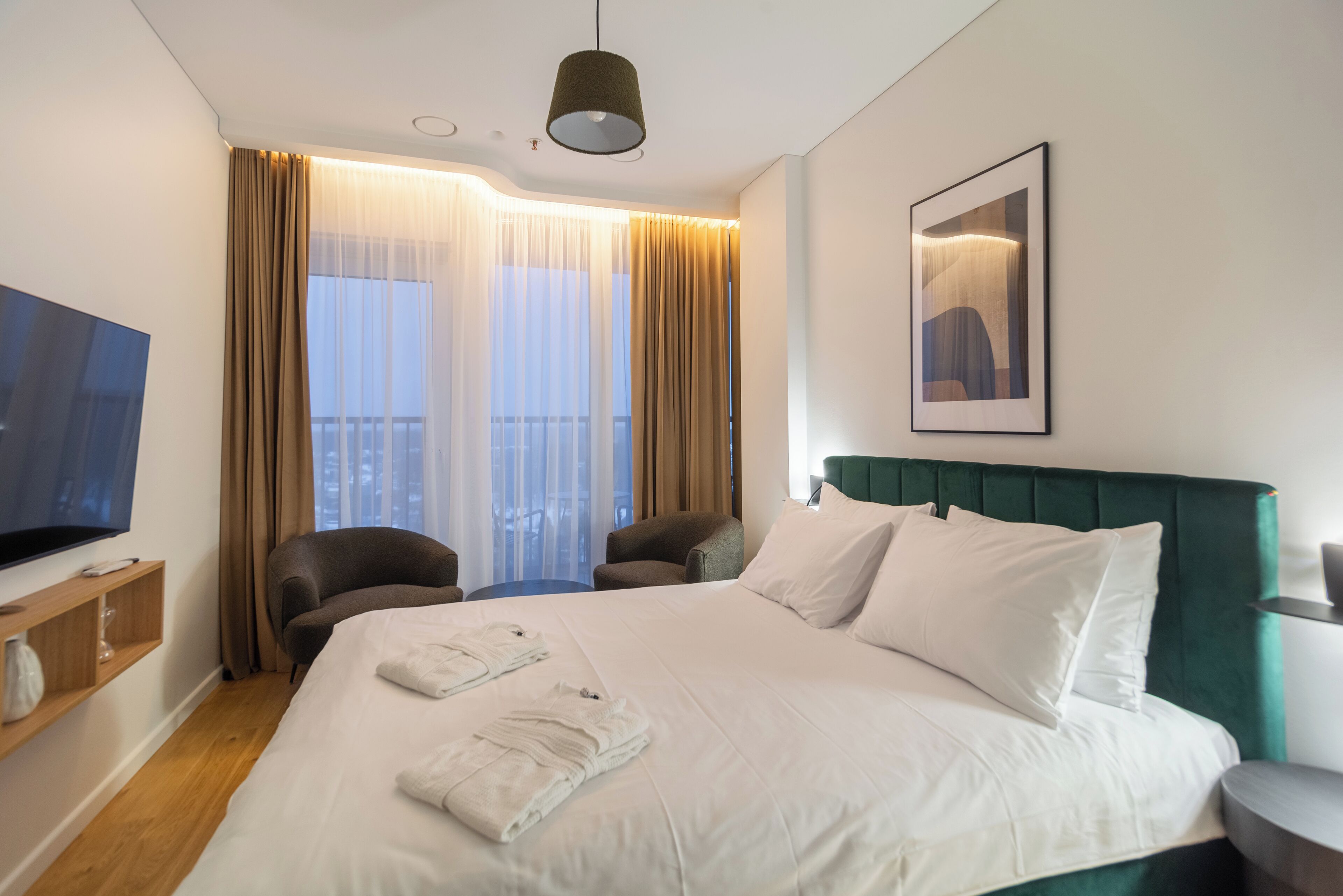 A hotel room with a large bed, two armchairs, a TV, and a painting on the wall.