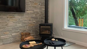 A modern living room with a stone wall, a black fireplace, a flat-screen TV, and a large window with a view of trees. - Oceanfront Views, Full Kitchen,  Wi-Fi ~ Where the Forest Meets the Ocean (Galiano Island)