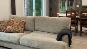A living room with a sofa, a dining table, and chairs. There is a window with a view of trees and a brick wall. - Oceanfront Views, Full Kitchen,  Wi-Fi ~ Where the Forest Meets the Ocean (Galiano Island)
