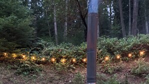 An outdoor seating area with two green chairs and a table, a wood-burning stove, and string lights in a forest setting. - Oceanfront Views, Full Kitchen,  Wi-Fi ~ Where the Forest Meets the Ocean (Galiano Island)