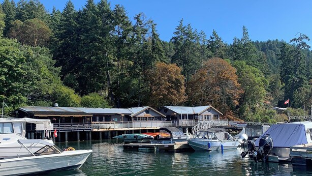 A marina with boats docked, a wooden boat in the foreground, and a building with a dock in the background. - Oceanfront Views, Full Kitchen,  Wi-Fi ~ Where the Forest Meets the Ocean (Galiano Island)