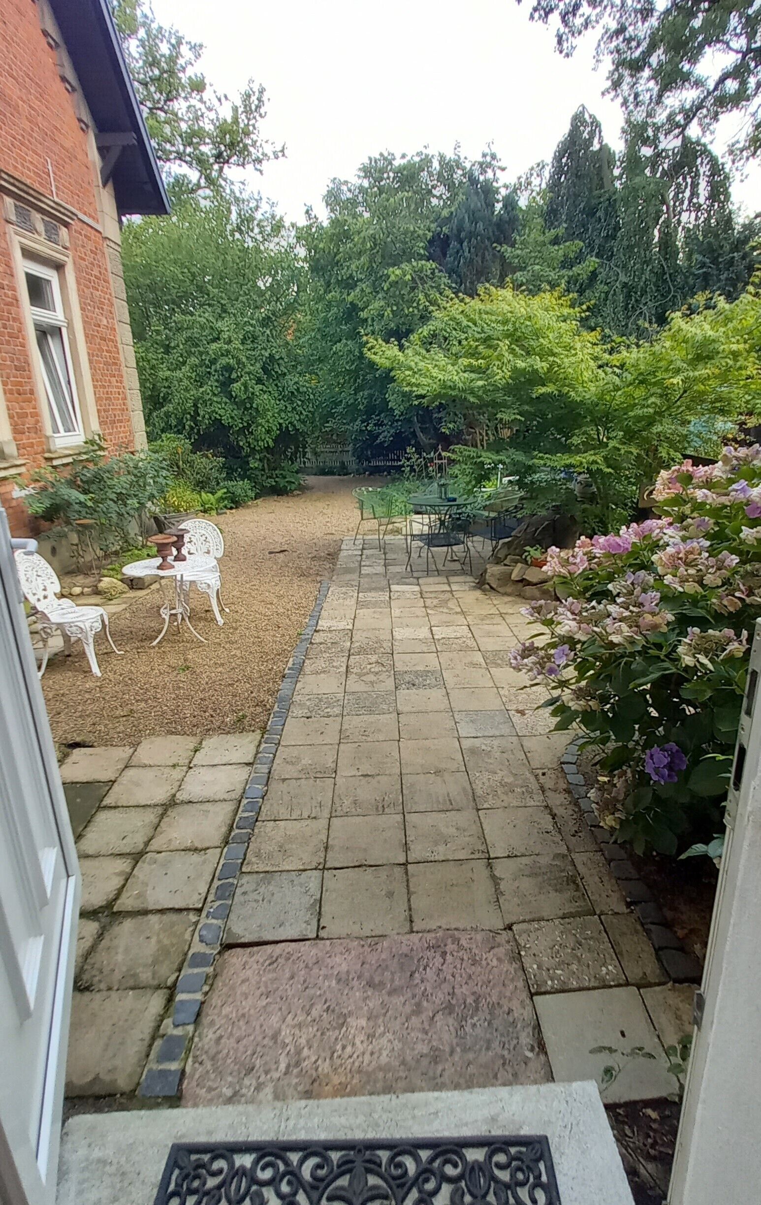 Eine gepflasterte Terrasse mit einem kleinen Tisch und Stühlen, umgeben von Grünpflanzen und einem Backsteinhaus.