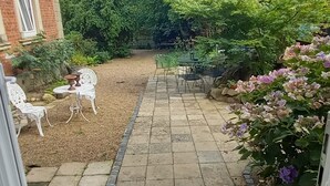 A paved patio with a small table and chairs, surrounded by greenery and a brick building.
