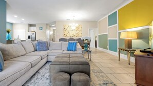 A modern living room with a sectional sofa, ottomans, a coffee table, and a dining area in the background. - 20 min to Disney , Epic , Star Wars , Gameroom ,Pool Luxe dw7 (Davenport)