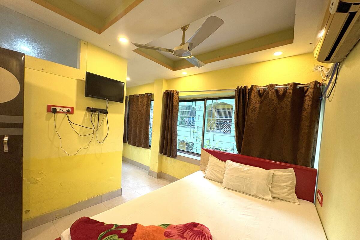 A hotel room with a bed, a ceiling fan, an air conditioner, a TV mounted on the wall, and a window with curtains.