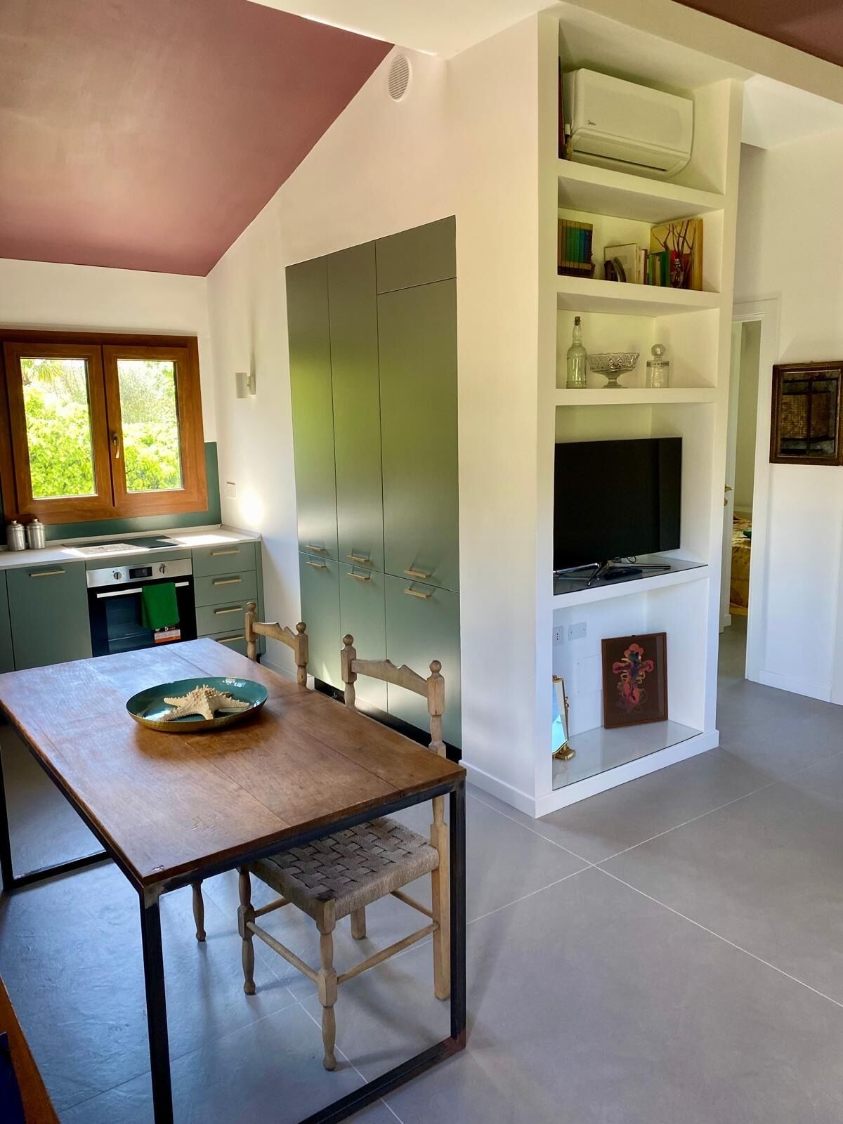 A modern kitchen with a dining area, featuring a wooden table and chairs, a built-in oven, and a TV on a white shelf.