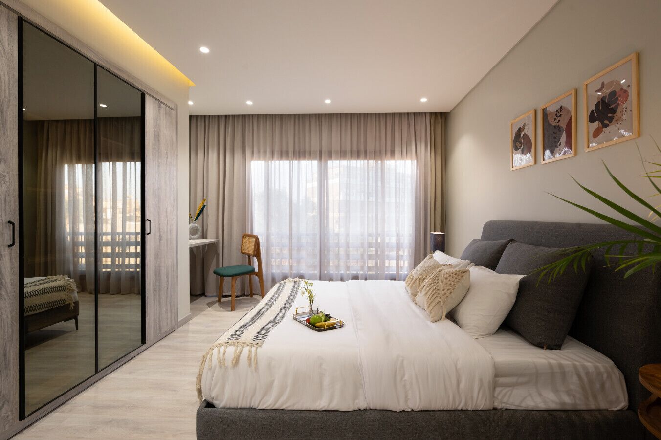 A modern bedroom with a large bed, a desk, and a view of the city through sheer curtains.