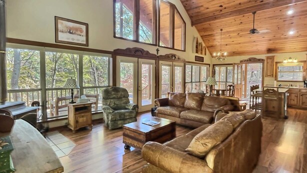 A spacious living room with a high ceiling, wooden beams, large windows, a leather sofa, a coffee table, and a dining area with a kitchen in the background. - Private Beaver Lake front Escape | 50 Acres (Rogers)