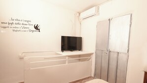Ein Zimmer mit weißer Wand, einem schwarzen Flachbildfernseher auf einer weißen Kommode, einem weißen Heizkörper und einer Tür mit grauem Rahmen.