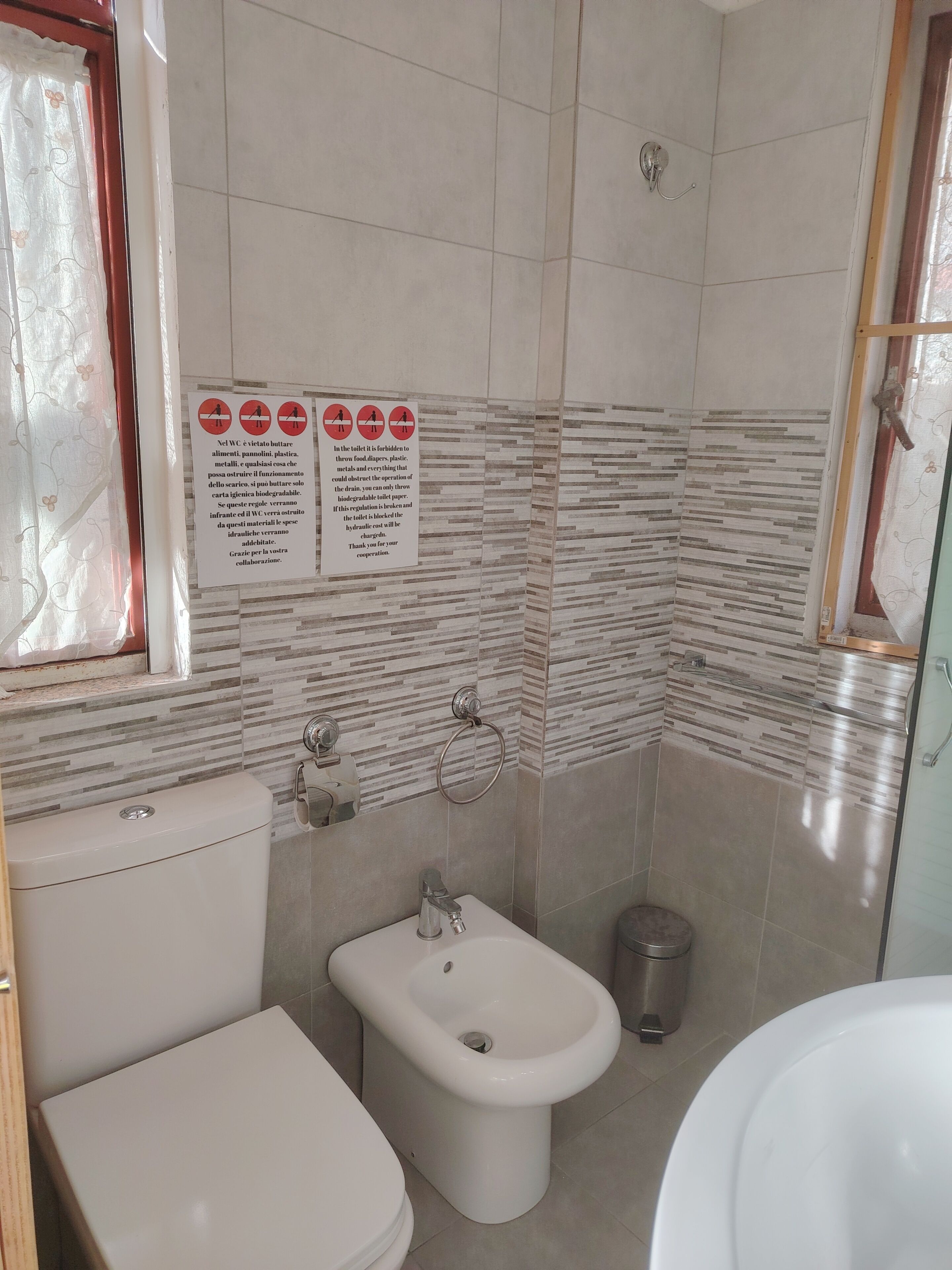 A bathroom with a toilet, bidet, and sink. There are tiled walls and a window with a curtain. A sign with instructions is visible on the wall.