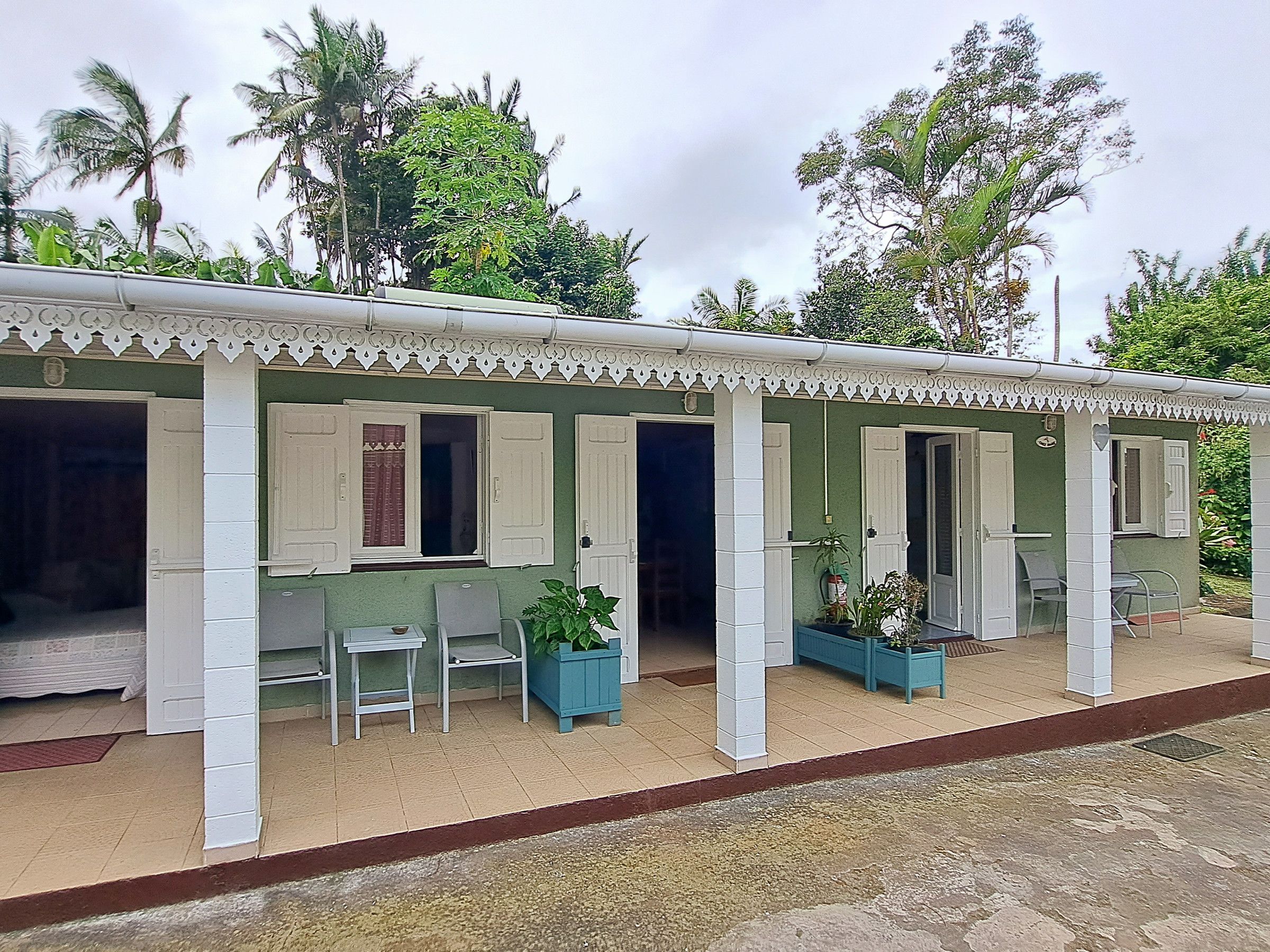 Un bâtiment à deux étages, doté d’une façade verte et de bordures blanches. Il possède de nombreuses fenêtres avec des volets blancs, et le rez-de-chaussée comprend un patio couvert avec des chaises et des jardinières.