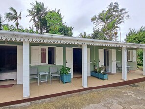 A two-story building with a green facade and white trim. There are multiple windows with white shutters, and the ground floor has a covered patio area with chairs and planters. - Authentic Creole Room in Les Avirons (Les Avirons)