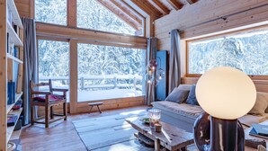 A cozy cabin interior with wooden walls and floors, large windows offering a snowy forest view, a wooden table with a lamp, and a comfortable seating area. - Luxury Chalet in Les Allues (Les Allues)