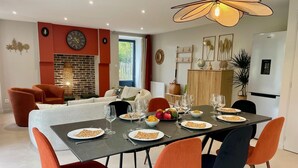 A dining area with a table set for six, surrounded by orange chairs, a modern chandelier, and a brick wall with a fireplace. - Cozy house in Montfarville, near Saint-Vaast-la-Hougue (Montfarville)
