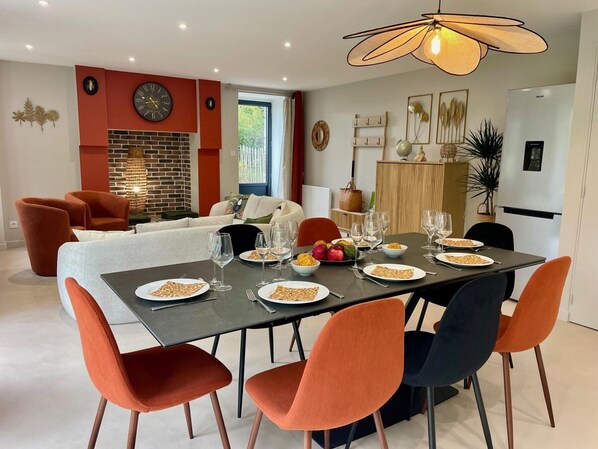 A dining area with a table set for six, surrounded by orange chairs, a modern chandelier, and a brick wall with a fireplace.