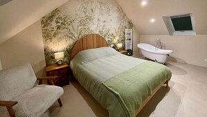 A bedroom with a sloped ceiling, a bed, a chair, a nightstand, a radiator, a bathtub, and a window. - Cozy house in Montfarville, near Saint-Vaast-la-Hougue (Montfarville)