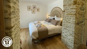 Ein Schlafzimmer mit Steinwand, Holzbalken, einem Bett mit Kissen und Decke sowie gerahmten Bildern an der Wand.