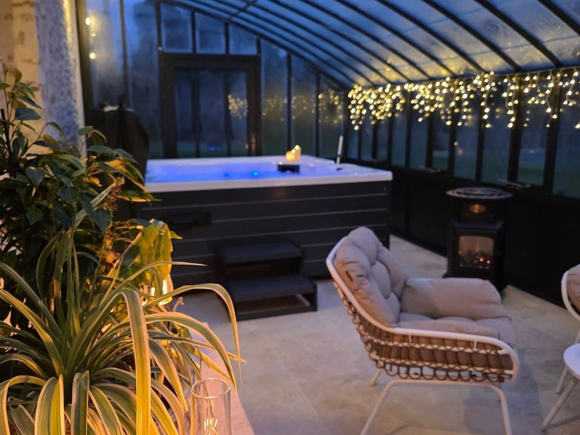 A greenhouse with a hot tub, wicker furniture, and string lights.