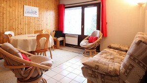 A cozy room with a dining table, chairs, a sofa, and a window with red curtains. - 3 Rooms in Carroz, 6 Pers, Terrace, Parking (Les Carroz d'Arâches)