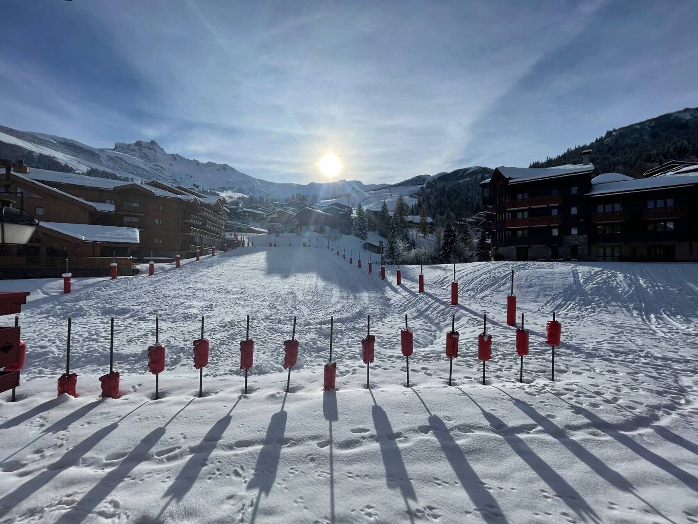 A snow-covered ski slope with red poles marking the path, surrounded by buildings and mountains.