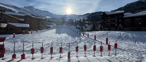 A snow-covered ski slope with red poles marking the path, surrounded by buildings and mountains.
