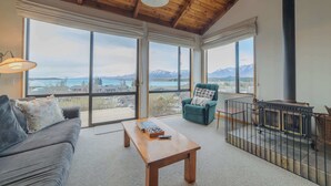 Un salon spacieux avec un plafond en bois, de grandes fenêtres offrant une vue sur les montagnes, un canapé confortable, une table basse et une cheminée.