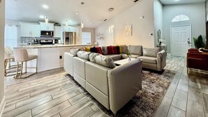Uma sala de estar espaçosa com um amplo sofá em formato de L, um espaço para refeições com uma cozinha ao fundo e uma área aconchegante com lareira.
