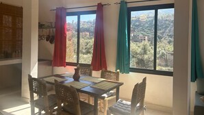 A dining area with a table and chairs, a view of a mountainous landscape through large windows, and a kitchen area with hanging pots. - Chez Yahia, in the heart of Ourika, apartment with panoramic view (Ourika)