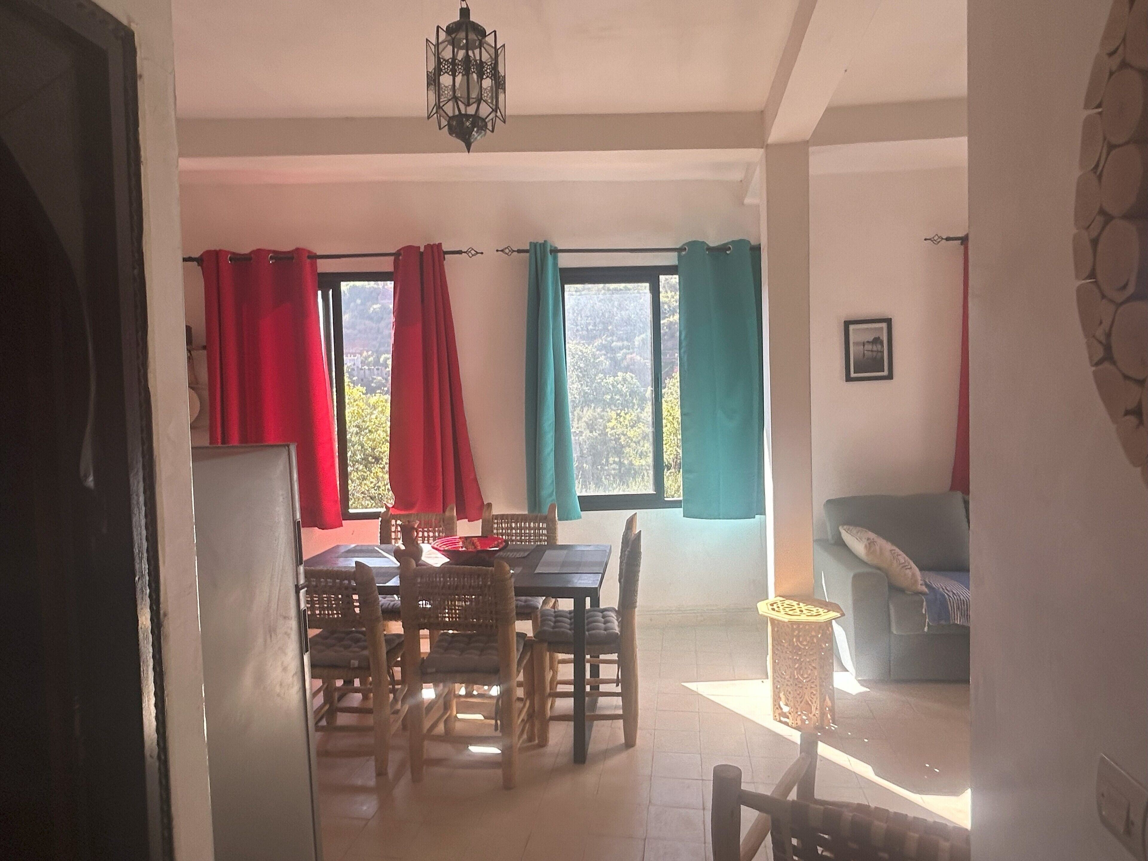 A dining area with a table and chairs, a hanging light fixture, and a view of the outdoors through windows with red and teal curtains.