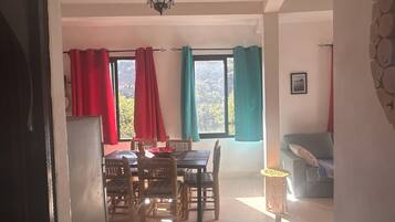 A dining area with a table and chairs, a hanging light fixture, and a view of the outdoors through windows with red and teal curtains.
