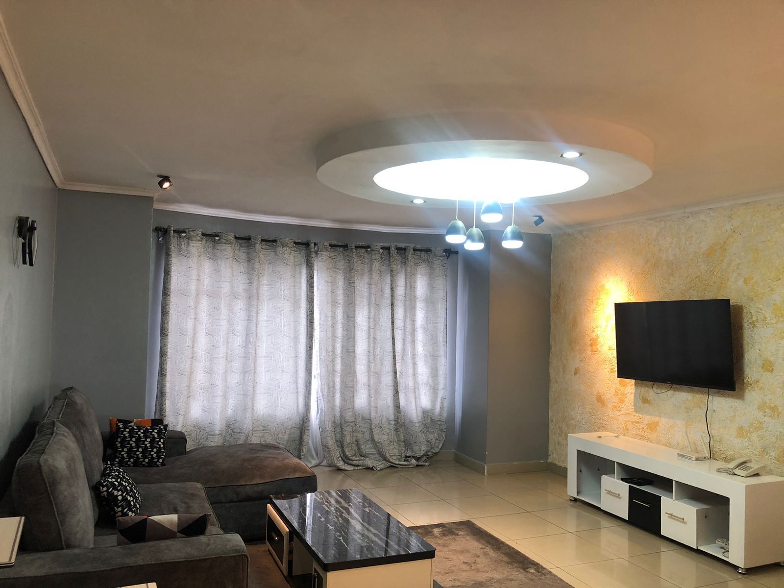 Una sala de estar con un sofá, una mesa de centro, un televisor sobre un soporte blanco y una lámpara en el techo.