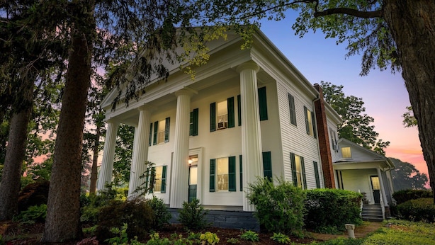 A two-story house with a porch, surrounded by trees and shrubs.