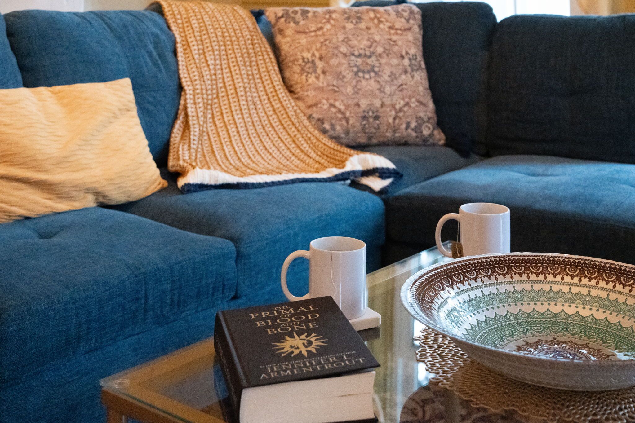En blå sofa med et mønstret plaid, en hvid kop, en bog med titlen "The Primal Blood Bone" og en dekorativ skål på et glasbord.
