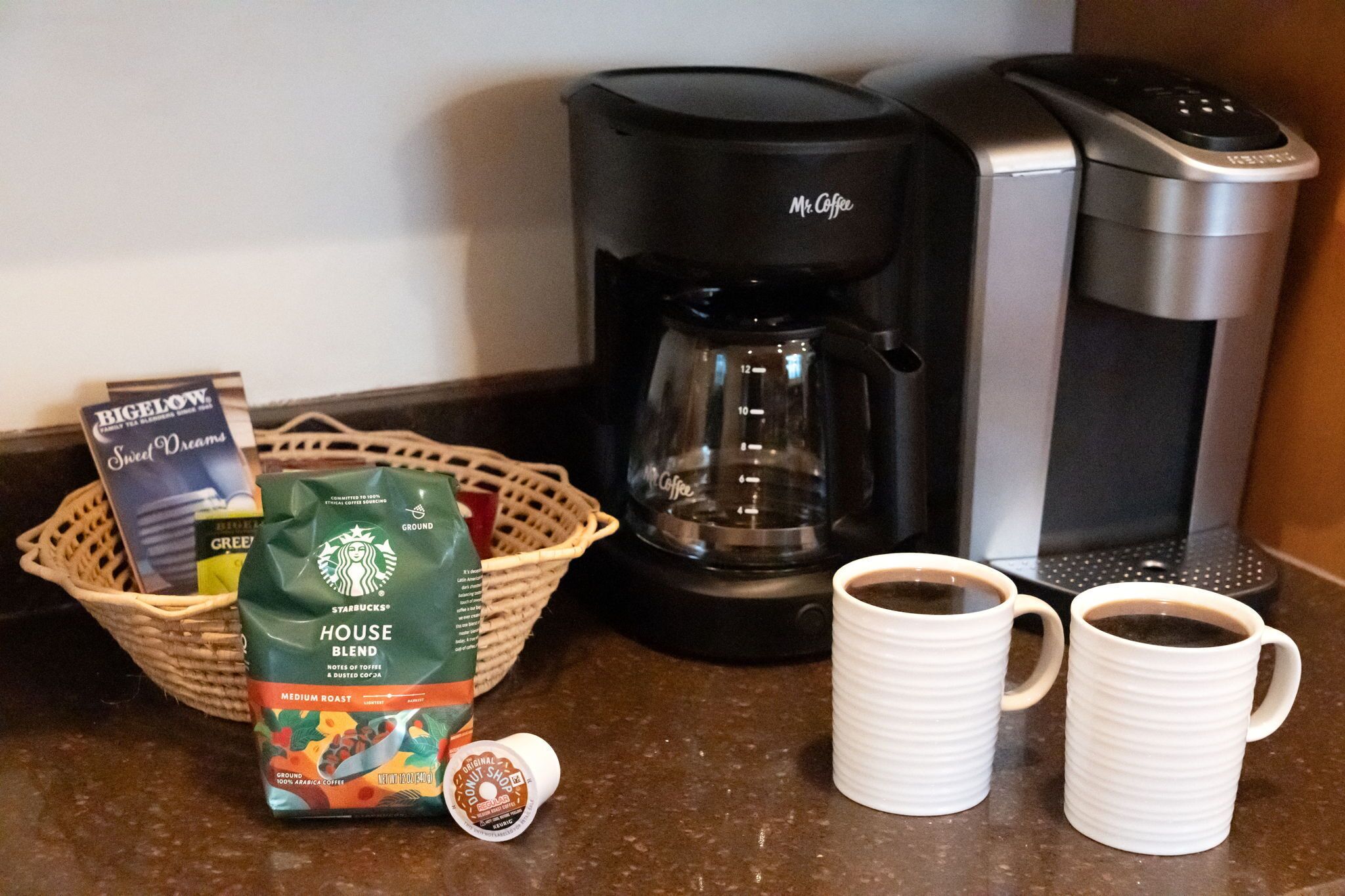 A coffee maker, two white mugs, a basket with coffee pods, and a bag of Starbucks House Blend coffee.