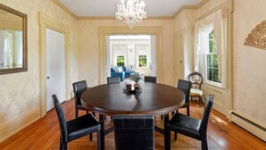 A formal dining room with a round table, six chairs, a chandelier, and a view to a living area with a blue sofa.