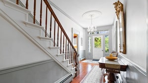 A hallway with a wooden staircase, a chandelier, a dining table with chairs, and a large mirror on the wall.