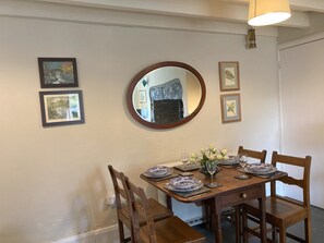 Ein Essbereich mit einem Holztisch, gedeckt für vier Personen, umgeben von Stühlen. An der Wand befinden sich gerahmte Bilder, über dem Tisch hängt ein runder Spiegel und auf dem Tisch steht ein kleiner Blumenstrauß.