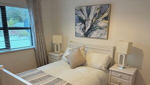 A bedroom with a white bed, two bedside tables, a nightstand, a window with blinds, and a painting on the wall.