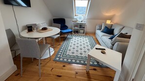 A compact living space with a round dining table, two chairs, a sofa, a small white table, and a patterned rug. A television is mounted on the wall, and a window provides a view of the sea.