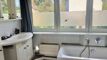 Een badkamer met een wit wastafelmeubel, spiegel en bad. Er is een raam met uitzicht op bomen en een hek.