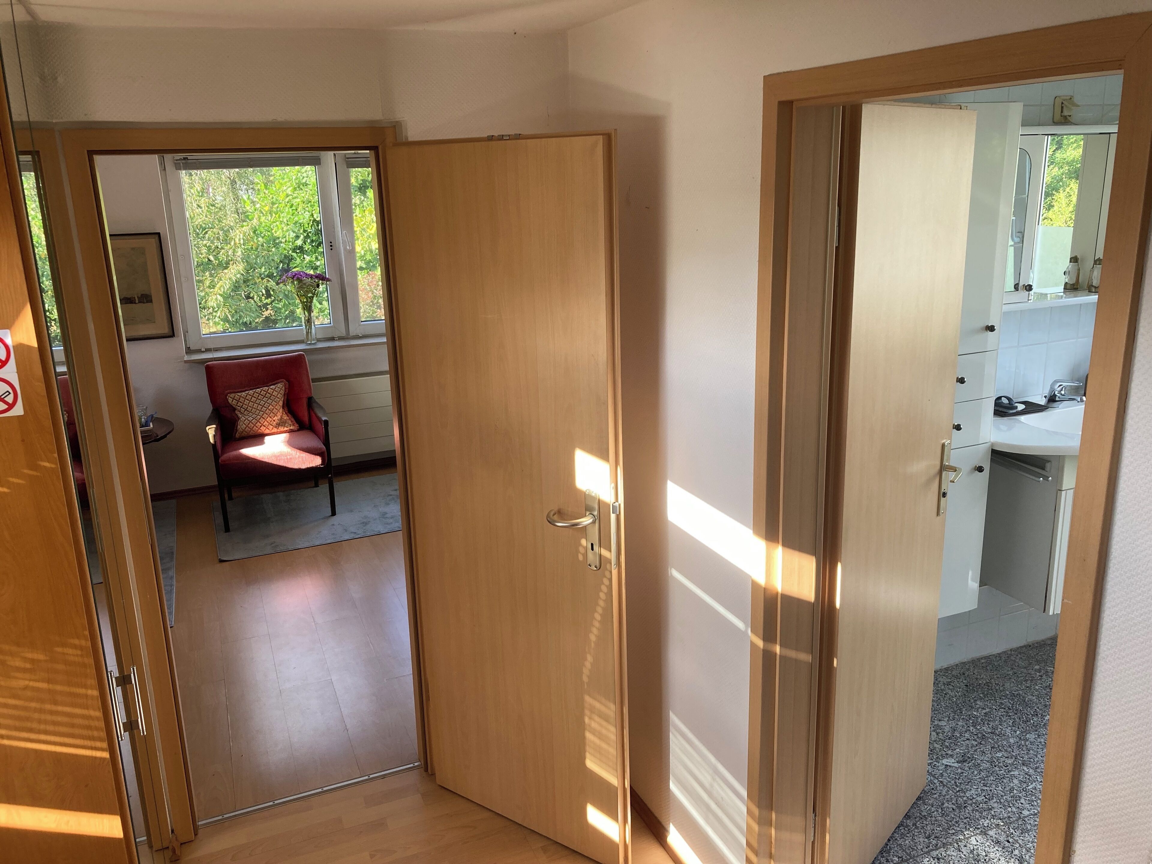 A room with a wooden door, a red chair, a small table, and a window with a view of greenery.