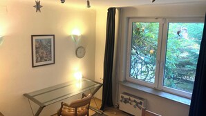 A room with a glass desk, a chair, a clock, a framed picture, and a window with a view of greenery. - Vacation apartment near Volkspark, arenas / Trabrennbahn /DESY (Hamburg)