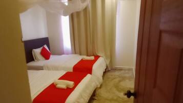 Habitación de hotel con dos camas, cada una con ropa de cama roja y blanca, una ventana grande con cortinas y una puerta de madera.