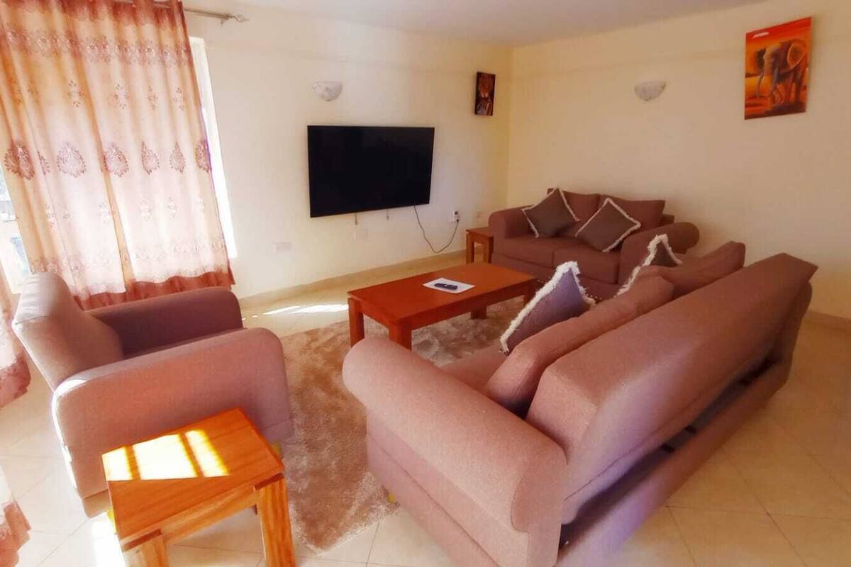 Una sala de estar con un juego de sofá, una mesa de centro, un televisor montado en la pared y una ventana con cortinas.