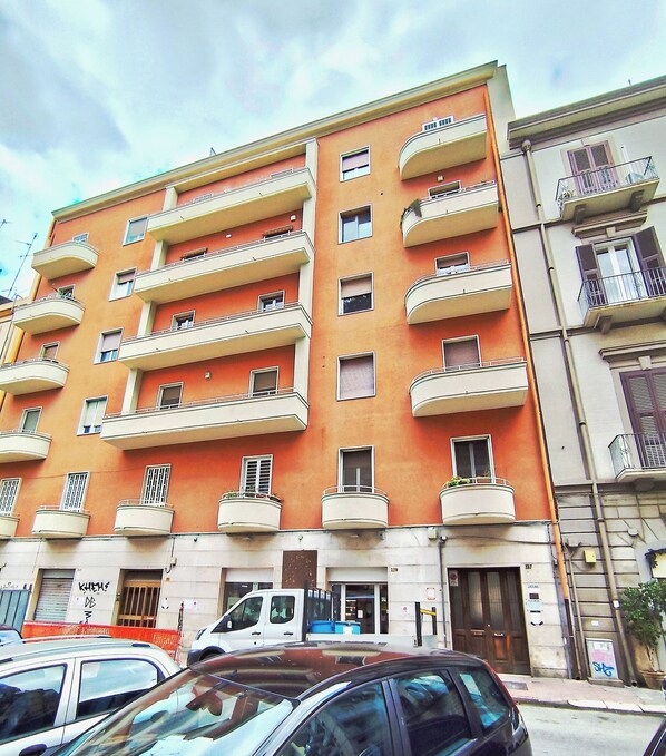 Un immeuble d’appartements à plusieurs étages, doté de balcons, avec un fourgon blanc garé devant et un pot de fleurs placé sur le trottoir.