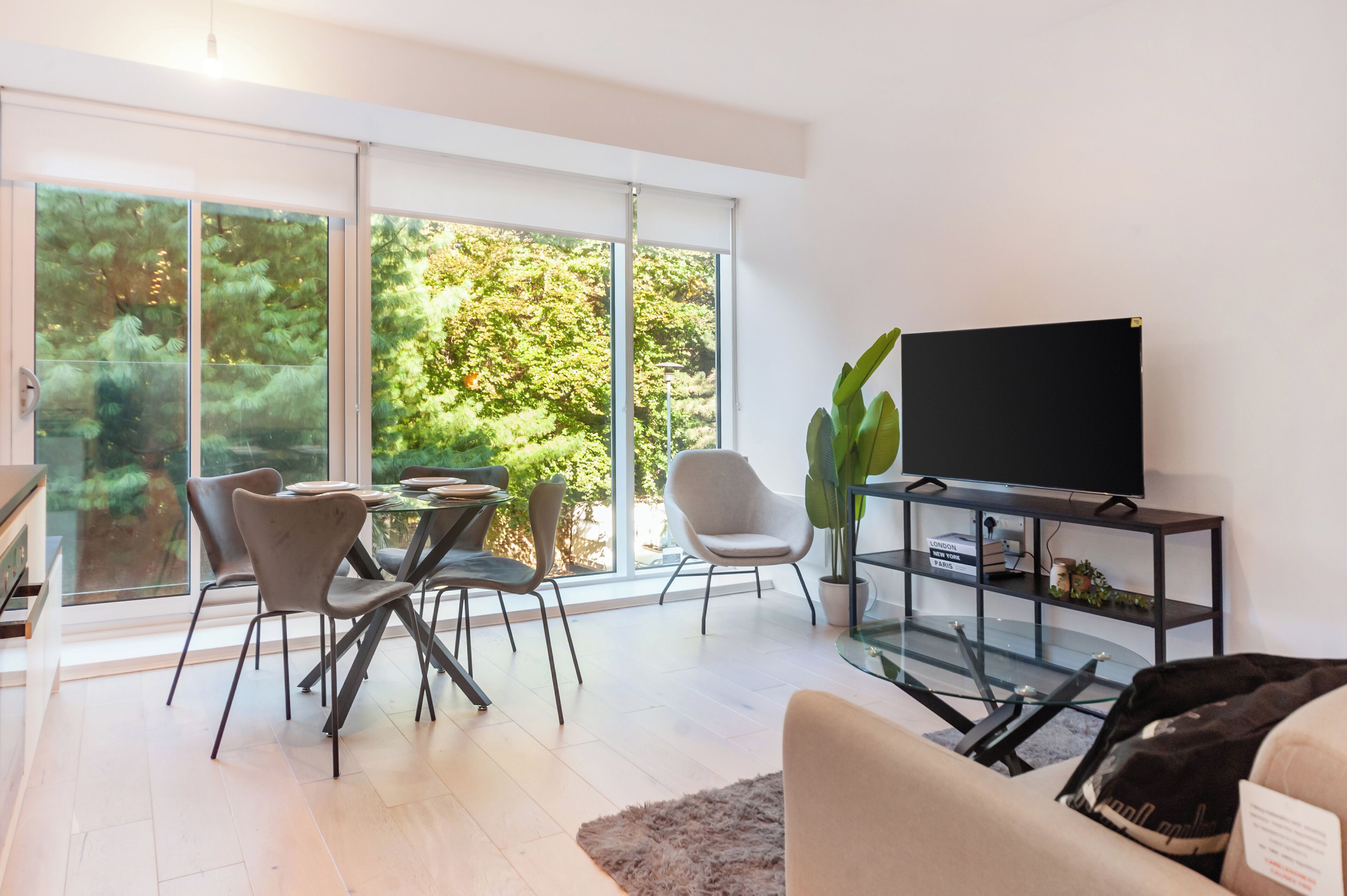 Ein modernes Wohnzimmer mit einem Glastisch, Stühlen, einer Couch, einem Fernseher auf einem schwarzen Lowboard und einem großen Fenster mit Blick auf Bäume.