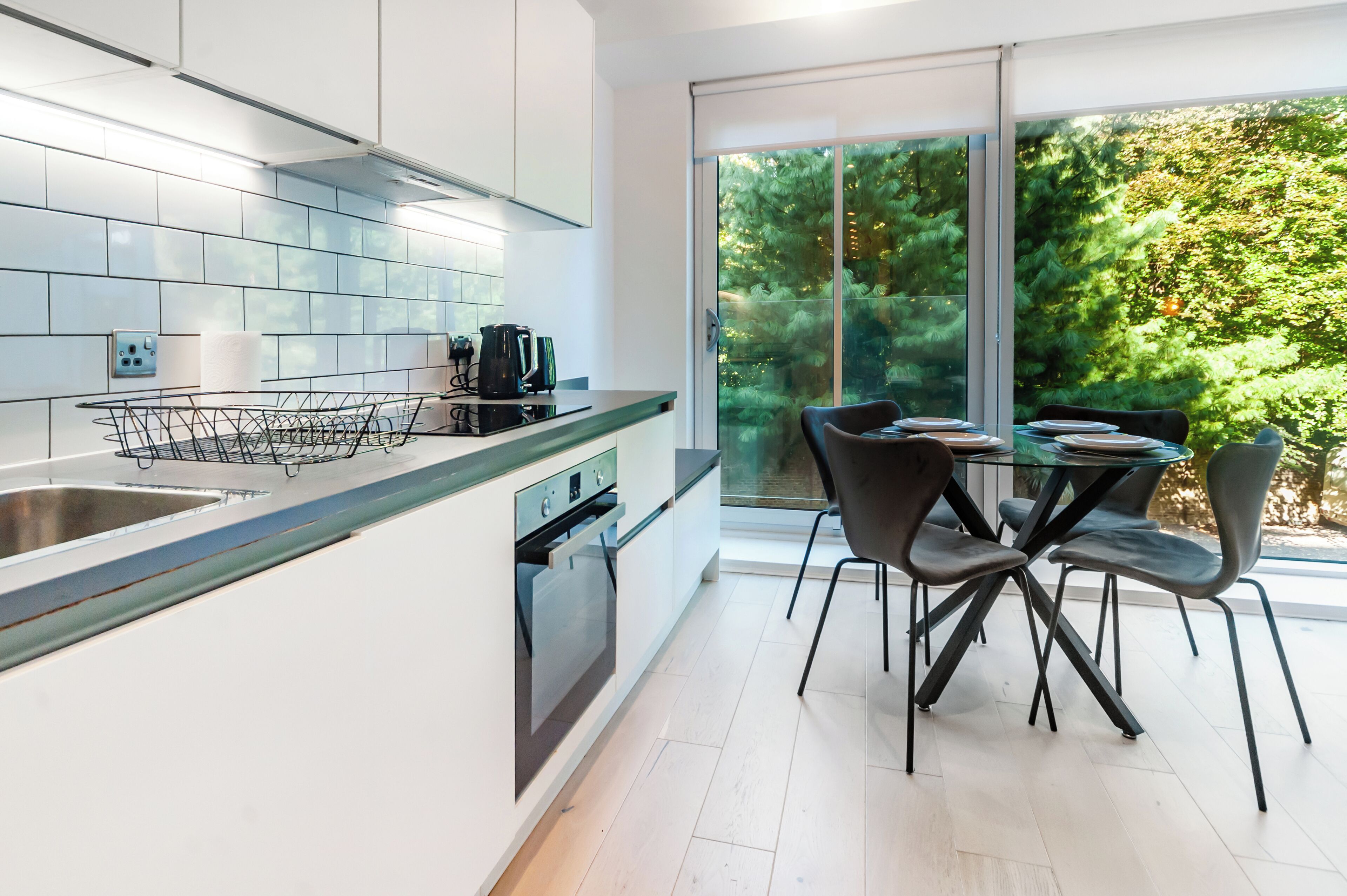 Una cucina moderna con mobili bianchi, un lavello in acciaio inossidabile e forno integrato. C'è un'area pranzo con tavolo in vetro e sedie nere.