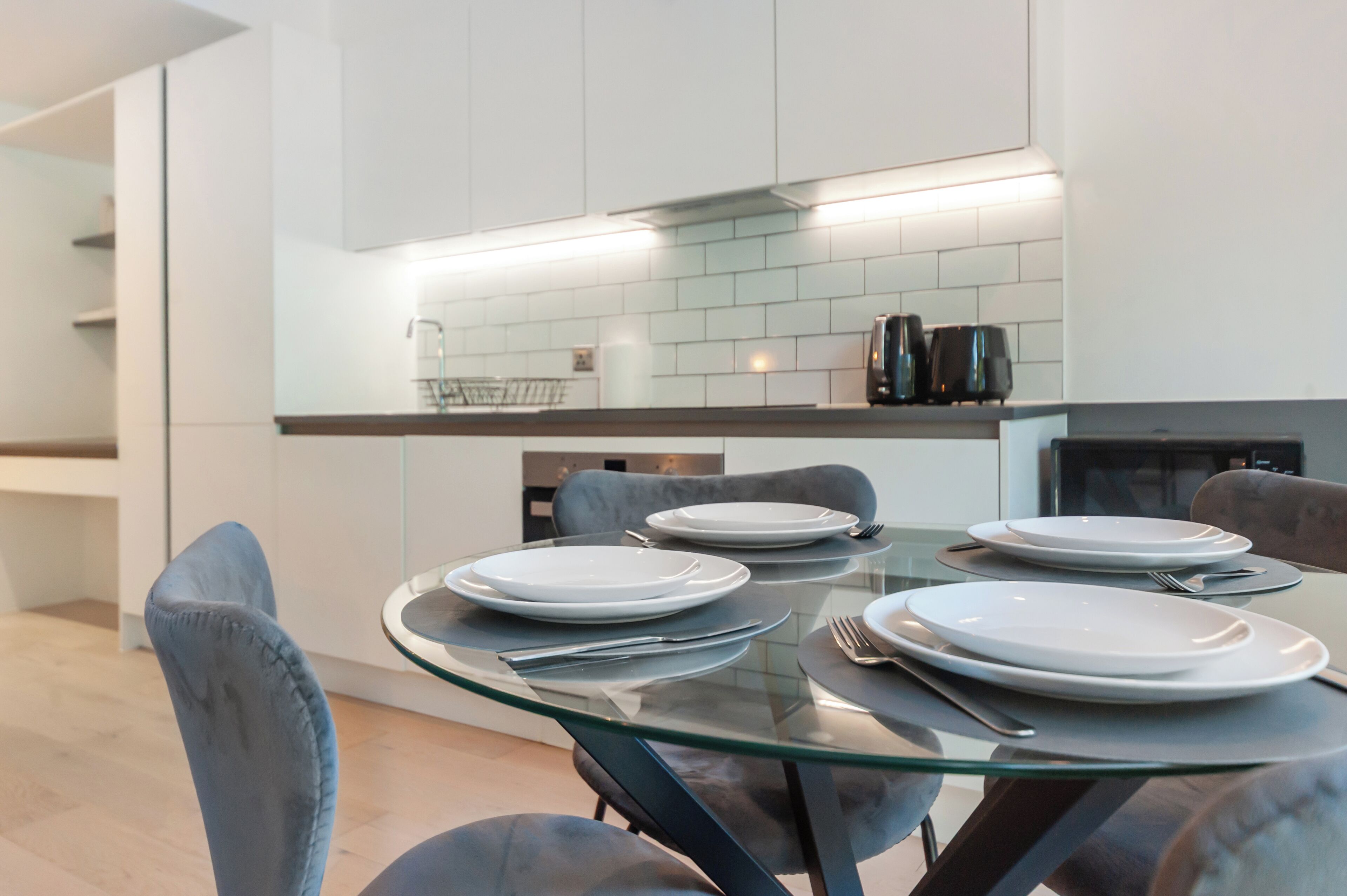 Een moderne keuken met een glazen eettafel, grijs gestoffeerde stoelen en witte kasten.